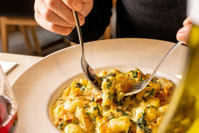 Hands holding fork and spoon to scoop up cheesy pasta.
