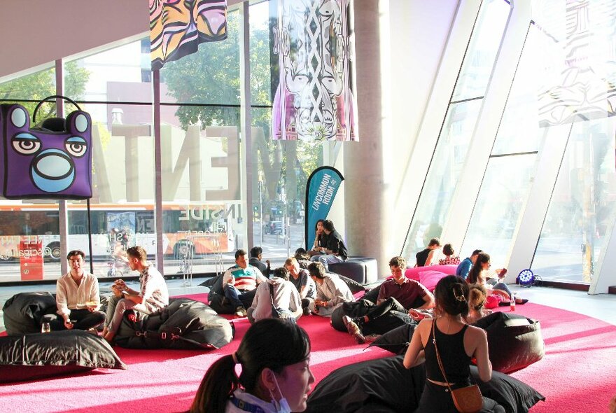People relaxing on bean bags in the Science Gallery, a large airy space with reddish/pink carpet, much glass and hanging displays.
