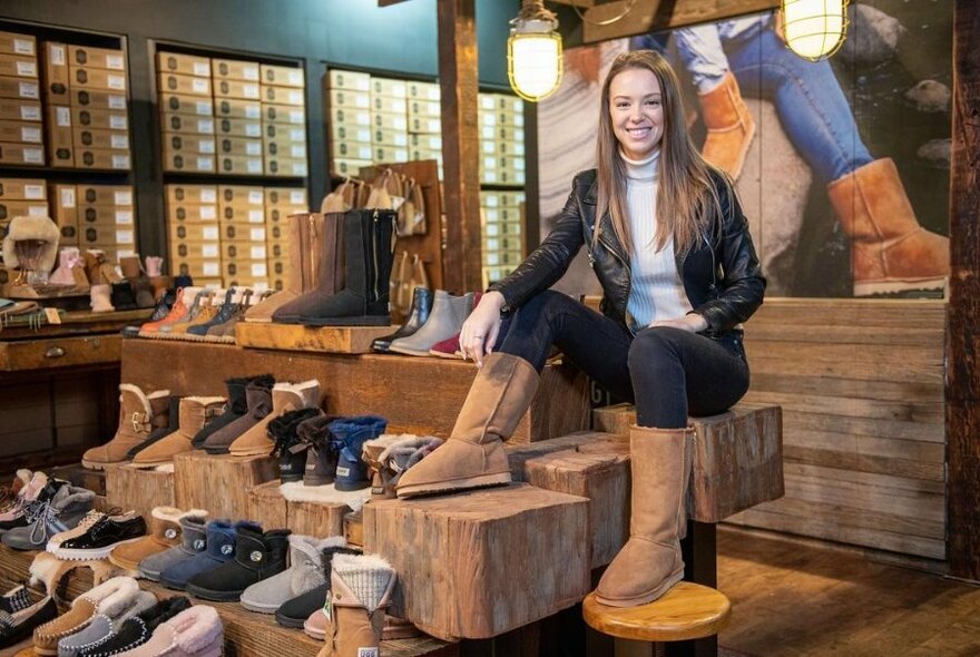 Person inside a store modelling Ugg boots of their feet, sitting on a display rack of many other Ugg boots and slippers.