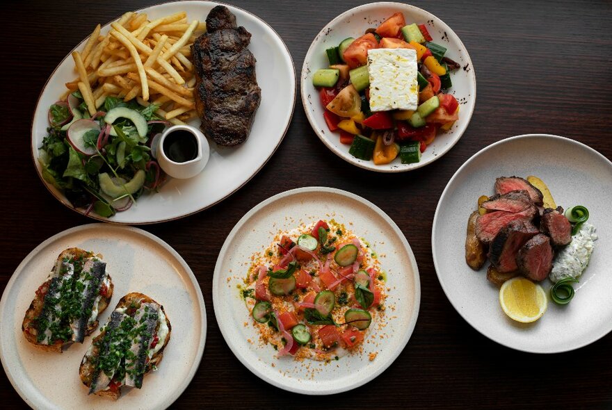 Looking down at meals that include a steak and chips, and a salad, served on white plates on a dark table surface.