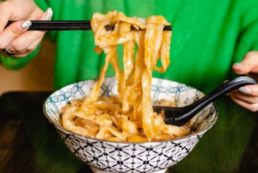Thick noodles held up with black chopsticks over blue and white bowl, green jumper at rear.