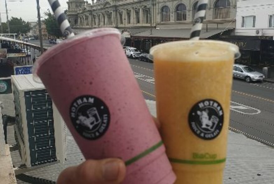 Two smoothies, one pink and one orange, in plastic cups held up against streetscape.