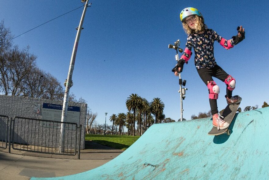 A girl skateboarder balanced at the top of a slide, wearing knee pads and a helmet.