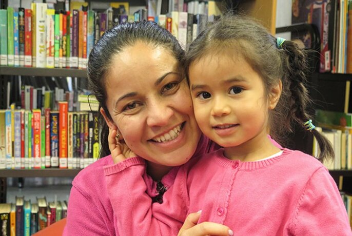 Smiling woman with her face pressed close to a preschool aged child's face, with shelves of library books behind them.