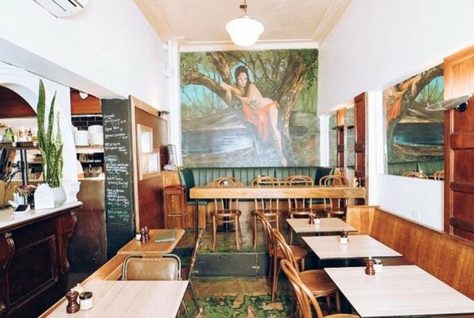 Interior of cafe showing tables and chairs and a large painting on the back wall.
