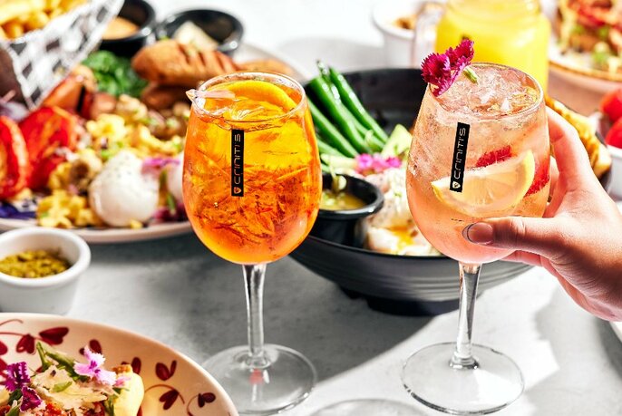 Two glasses of spritz on a table, with plates of food visible behind. 