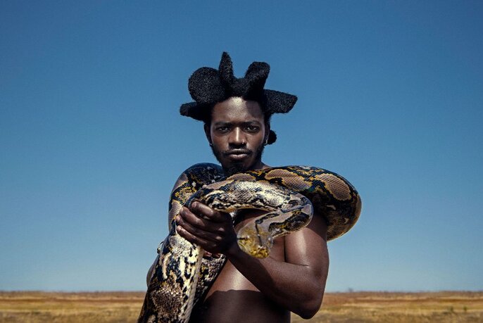 Singer and performer Desire Marea, standing in a desert landscape against a blu sky, and with a large python snake coiled around his neck and naked torso.