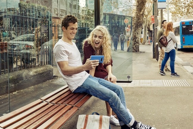Young man and woman sitting at a bus shelter and laughing while viewing something on the man's phone.