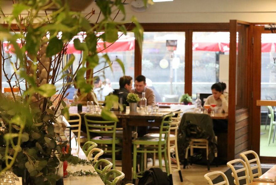 Restaurant interior with central floral arrangement and peopled seated at tables beside a window.