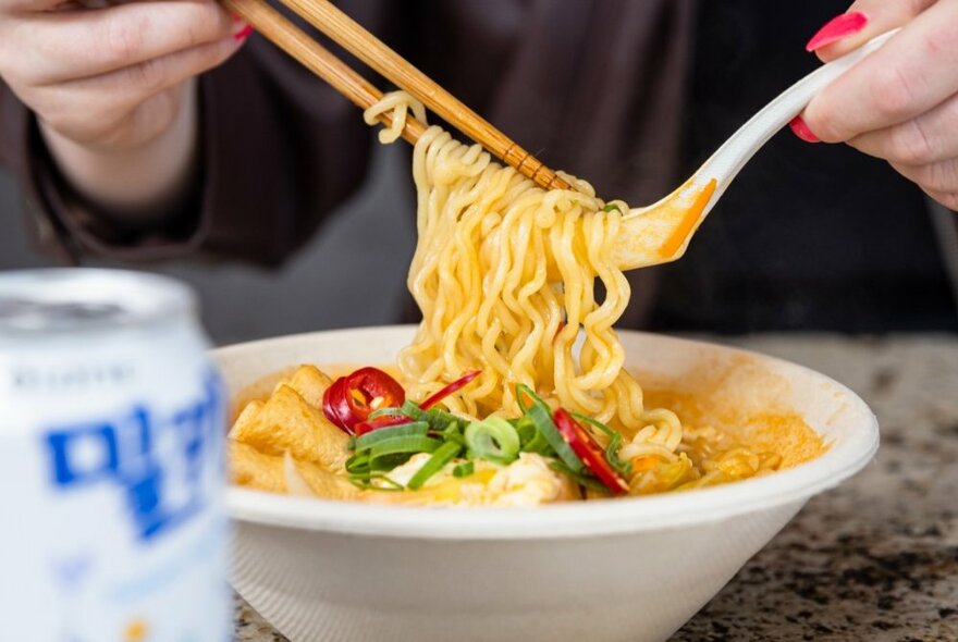 Hands using chopsticks and a spoon dipping into a bowl of noodles and vegetables, with a blurred can of drink in the foreground.