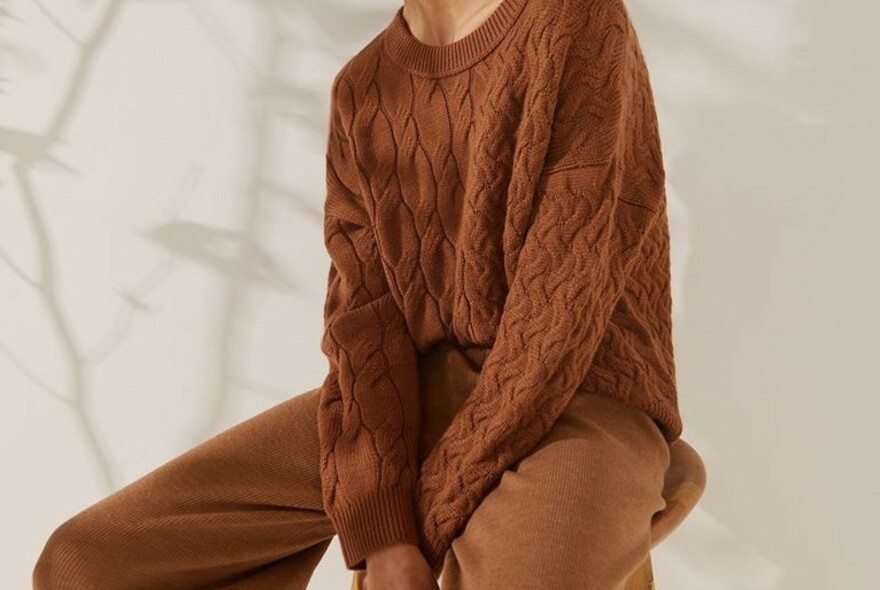 Model seated with arms folded wearing chunky light brown knitted sweater.