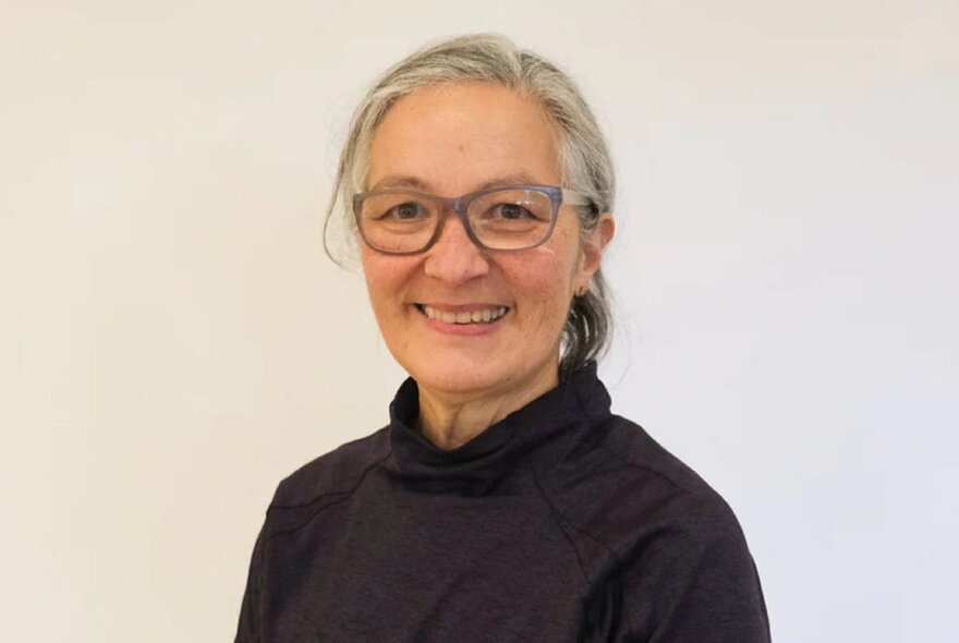 Female yoga practitioner smiling with glasses, grey hair and dark polo neck top.