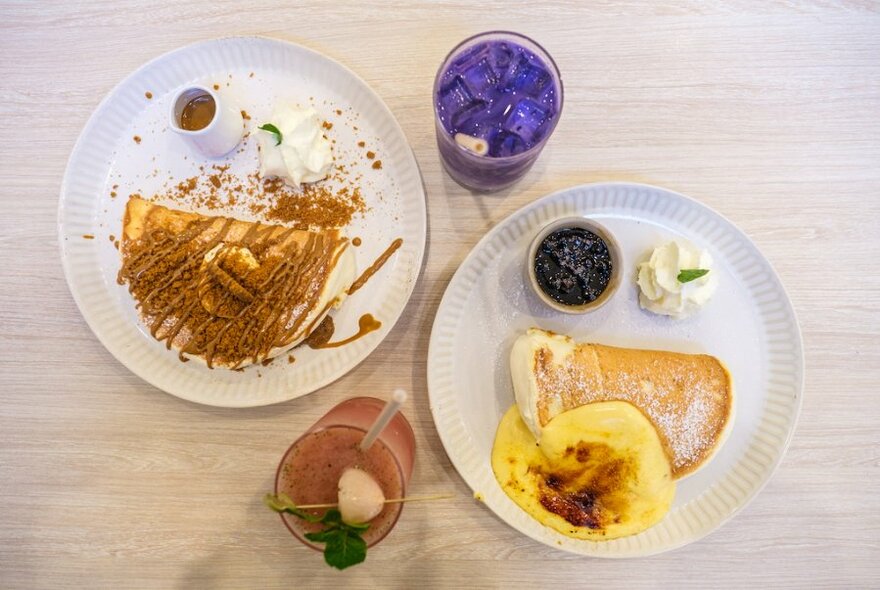 Looking down on a table with two plates of soufflé pancakes and two coloured drinks. 