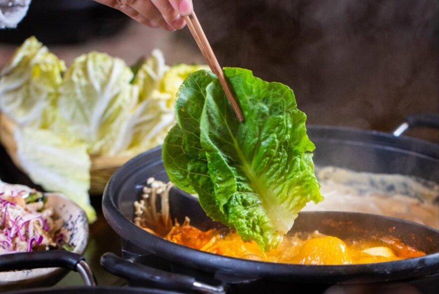 A hand holding chopsticks dipping a lettuce leaf into a steaming pot.