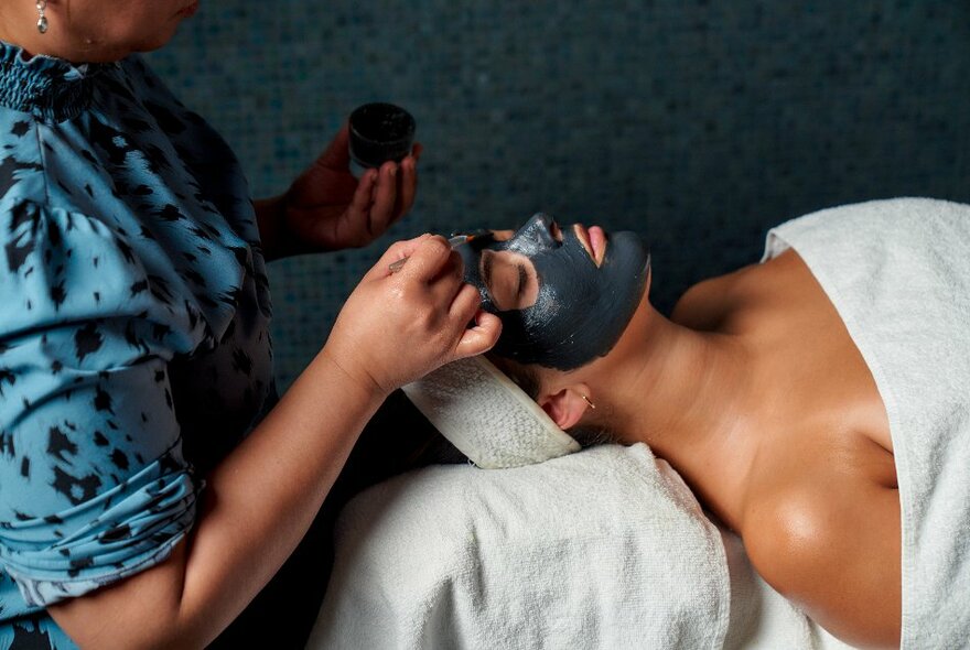 A therapist applying a dark face mask on a customer lying on a table, their head and body wrapped in white towels.