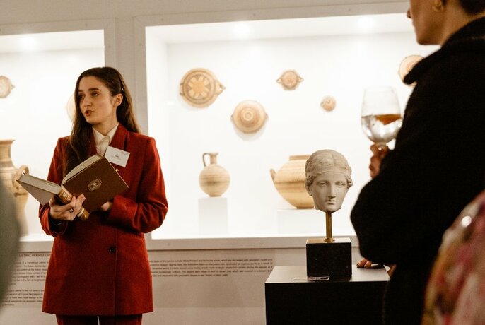 Woman with long dark hair and red jacket, holding hardback book, posed as if addressing group, lit cabinets with antiquities behind.