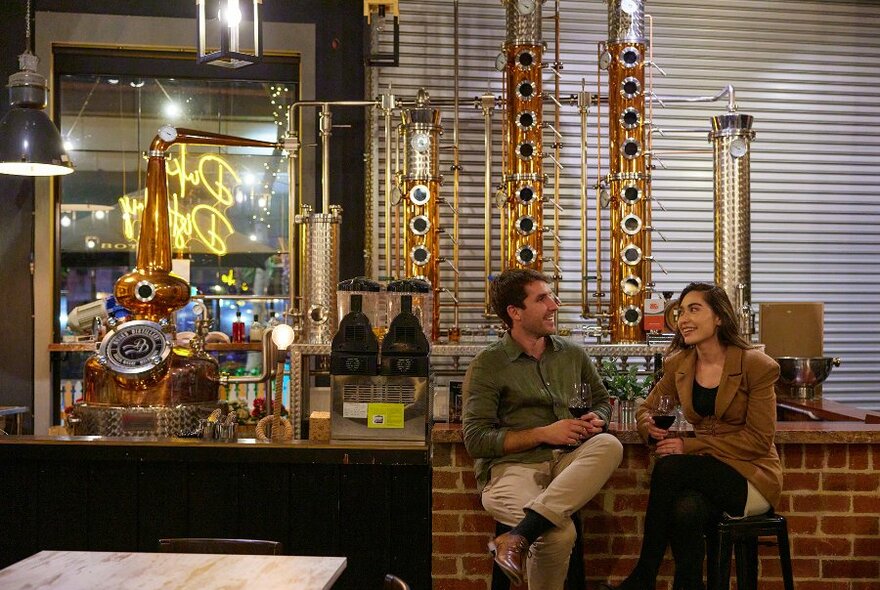 A couple sitting at a bar in front of a large copper still. 