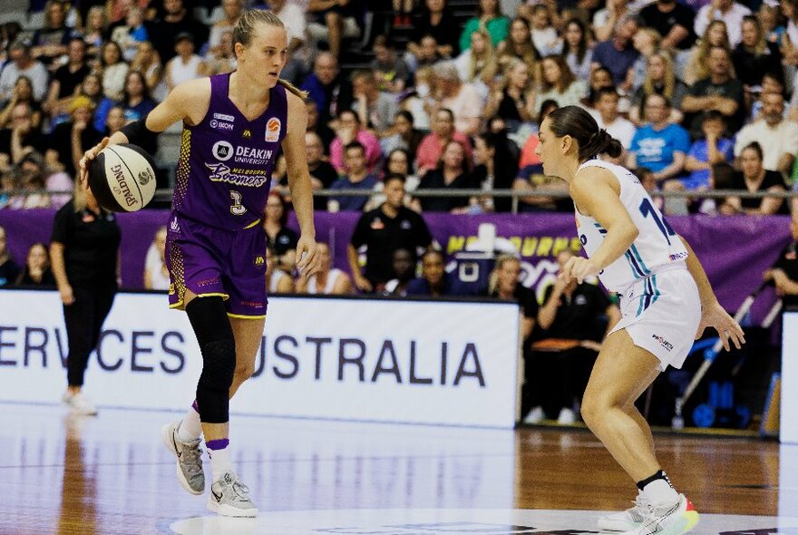 Women playing basketball on a court, one person dribbling the ball by bouncing it on the ground, with a large stadium crowd in the background.