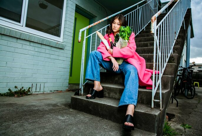 Model wearing a pink coat and jeans sitting looking glum on the outside steps of a dingy block of flats.