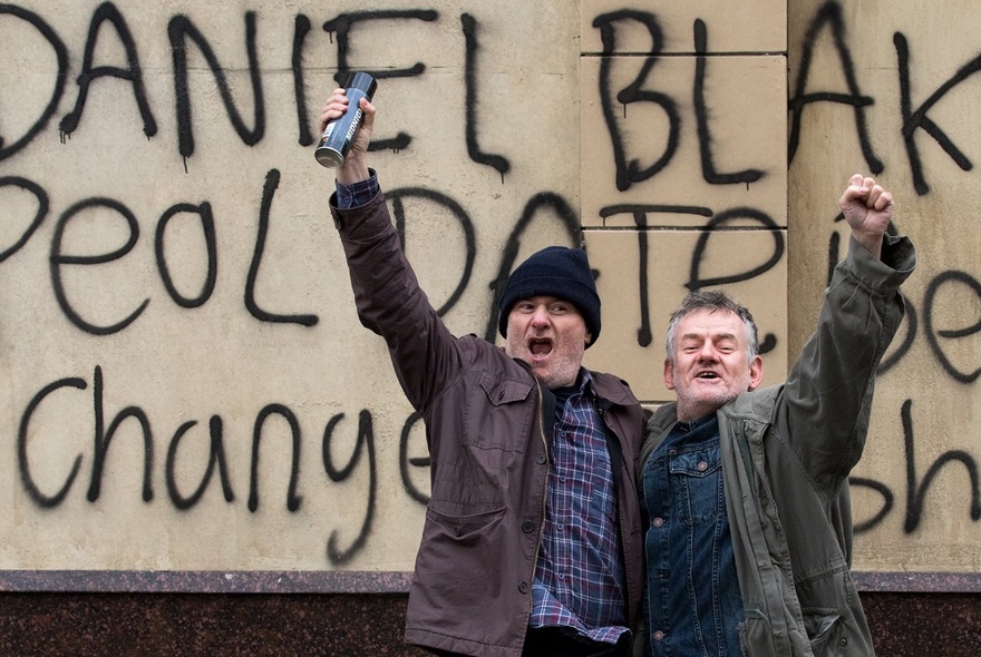 A still from a Ken Loach film featuring two men with arms raised in celebration, in front of political graffiti on a signboard.