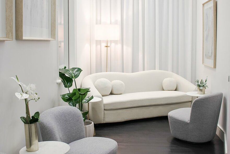 Room interior with a white couch, soft grey armchairs, white walls, white curtains and green plants.