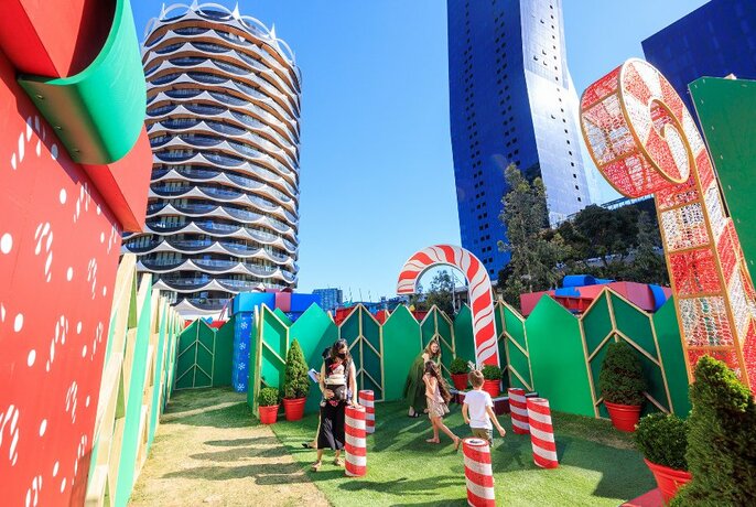 Parents and children play on a grassy area surrounded by tall green walls and candy canes, with city buildings in the background.