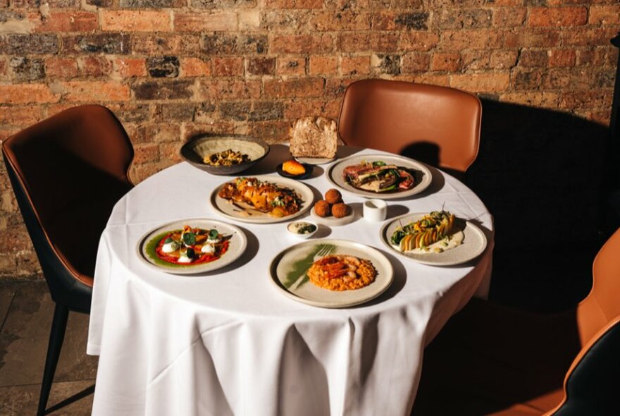 Outdoor tables with white cloth, against brick wall, full of plates of pasta. 
