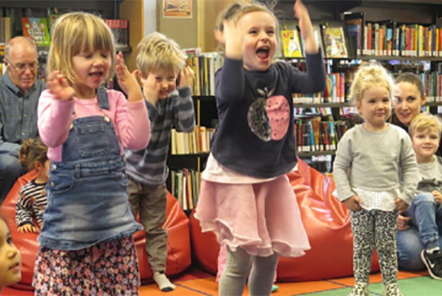 Pre-school aged children jumping up and down in a library setting, guardians and bookshelves behind them.