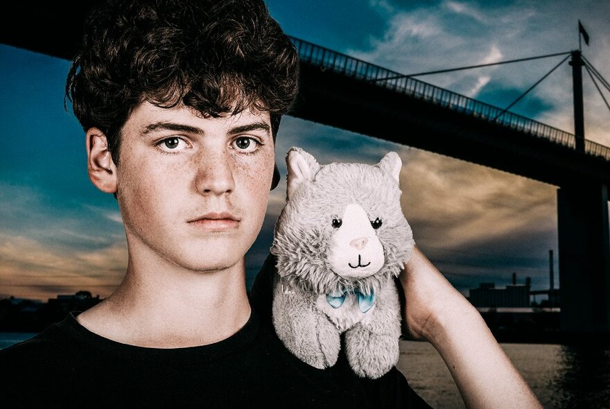 A teenage boy looking directly at the camera holding a grey soft toy on his shoulder, in front of a backdrop of a dusky sky and silhouette of a bridge.