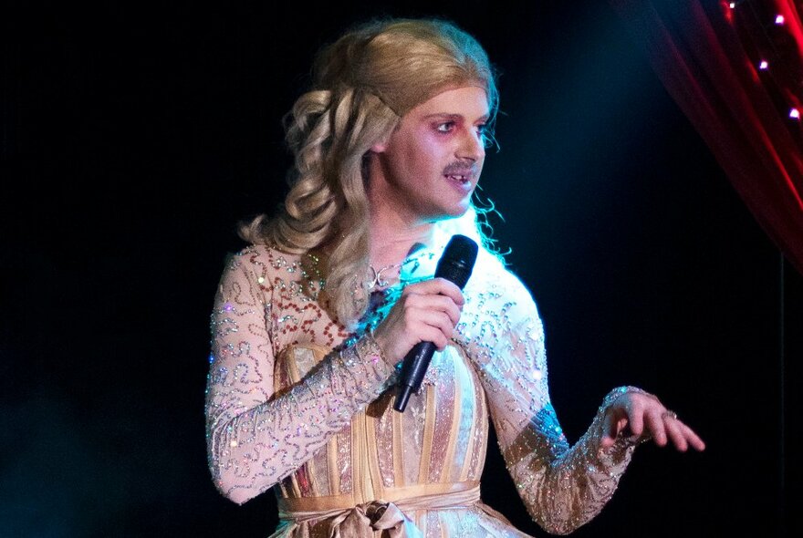 A performer in costume on stage, wearing a blonde wig and talking into a microphone.