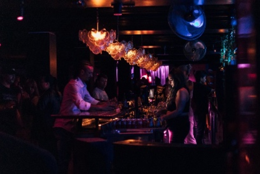 Dimly lit nightclub bar, with patrons seated on one side of the bar counter, and a person serving drinks on the other side.