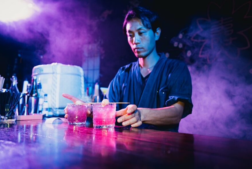 A bartender with pink drinks at a wooden bar with atmospheric smoke.