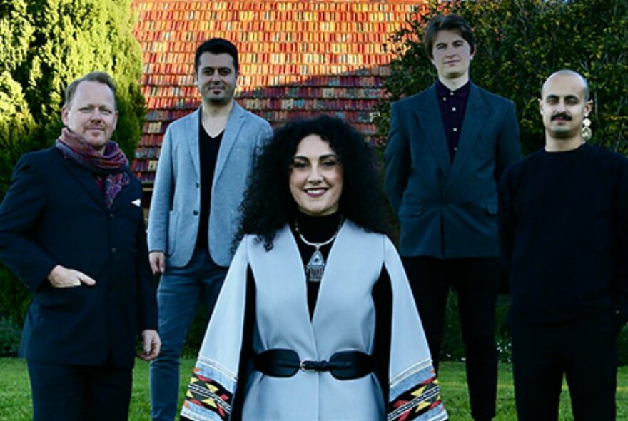 Five band members arranged with a woman in the front in a pale blue jacket. 