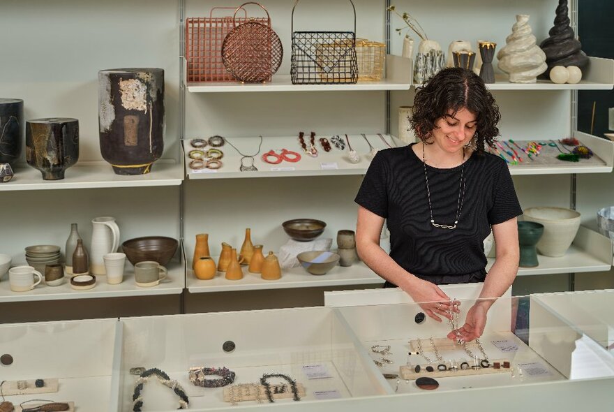 A person gently lifting a jewellery item from a glass display cabinet, shelves of art and craft objects on display behind her.