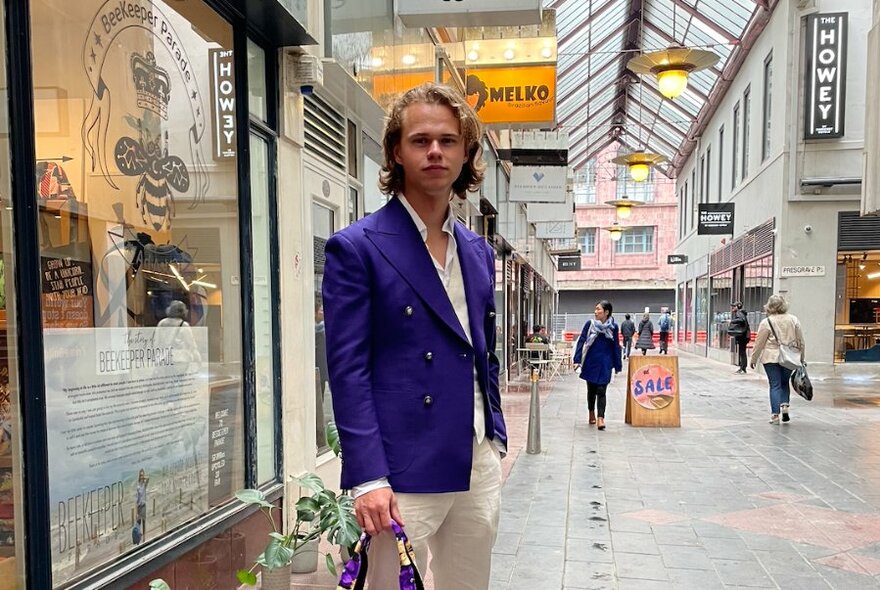 Person in a purple jacket and light-coloured trousers, standing outside a retail space in a shopping arcade.