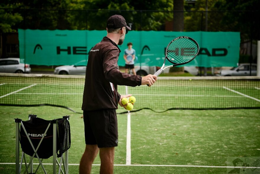 Rear view of a person standing on a grass tennis court about to hit a ball with a tennis racquet to a person standing at the opposite end of the court.