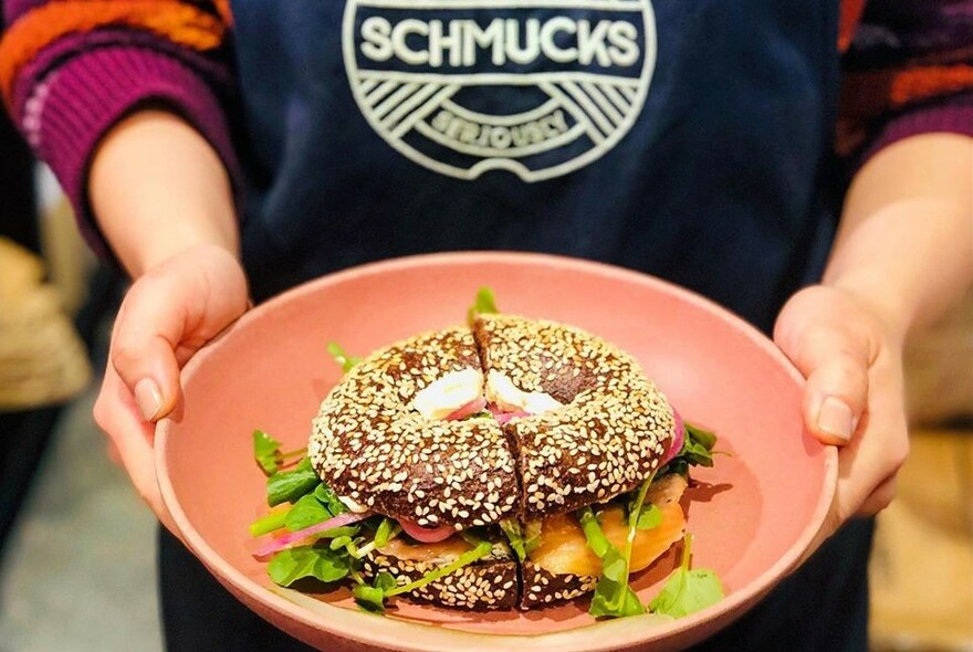 Waiter wearing Schmucks apron holding a pink bowl with a sesame seed salad bagel.