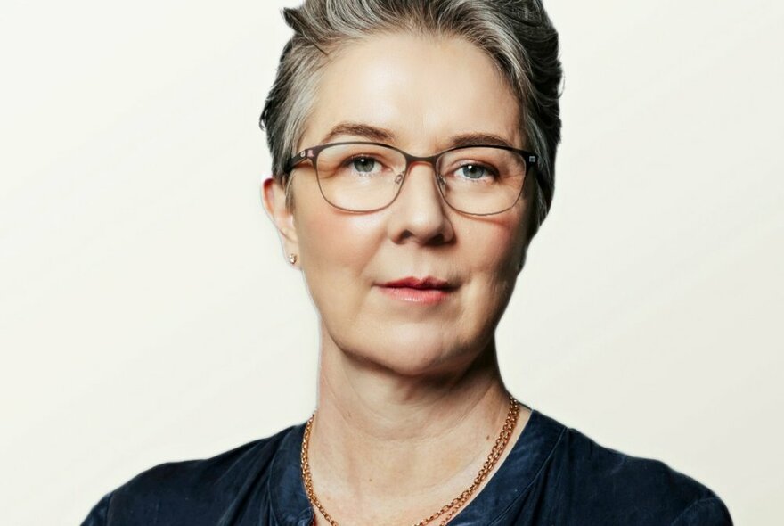 Head and shoulders of a women looking intently at the camera, short silver grey hair, wearing glasses and posed against a white background.