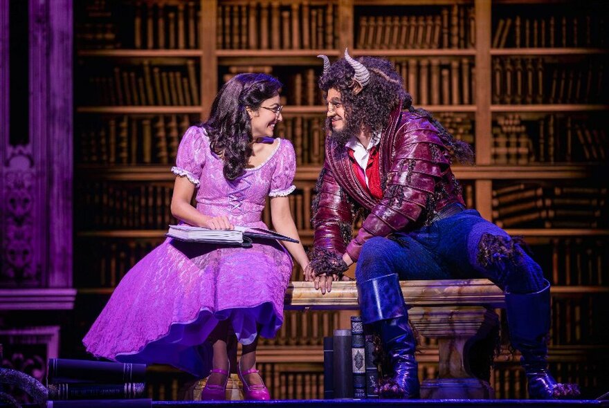 Two cast members from the musical Beauty and the Beast - the beast and the beauty - on stage, sitting on a bench with library shelves as a backdrop, looking at each other and smiling.