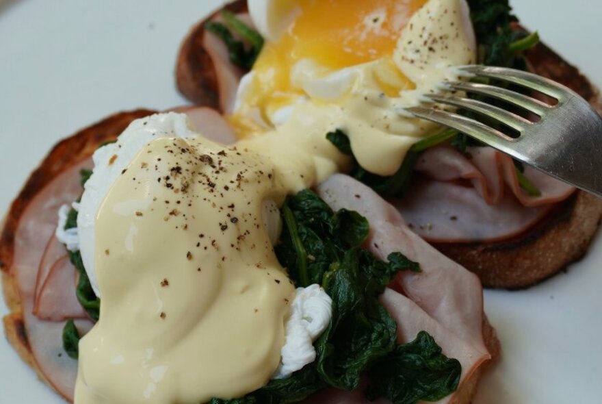 A fork piercing a poached egg on a pile of spinach and ham on toast.