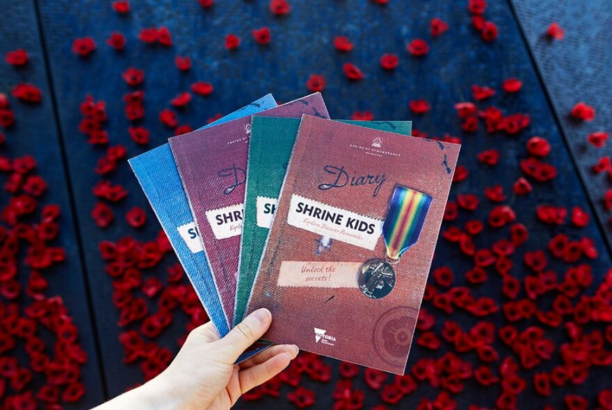 A hand holding four booklets about the Shrine in front of a wall of poppies.