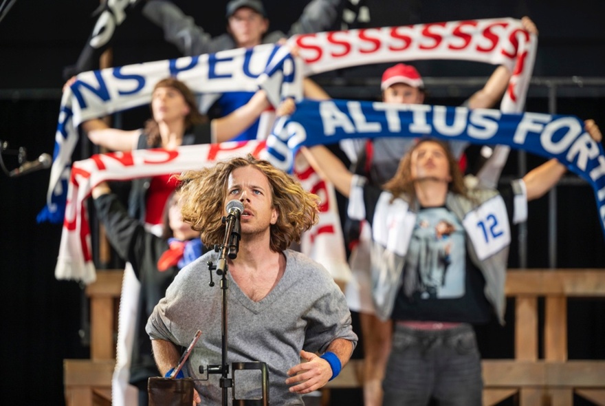 A person singing in front of a microphone in the foreground, behind them a small group of people waving football scarfs in the air above their heads.