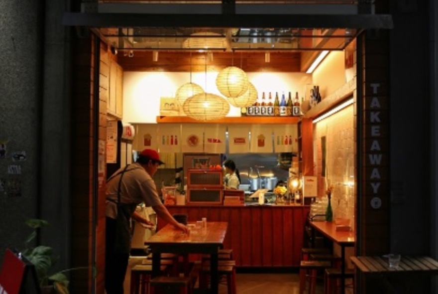 View from a laneway into a warmly lit and cosy interior of a cafe, a man setting a table inside and a view of the kitchen at the rear of the cafe. 