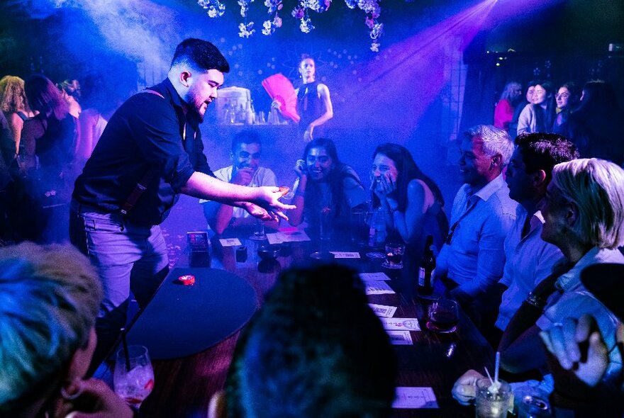 A performer with cards in front of seated patrons in a smoky dark bar.
