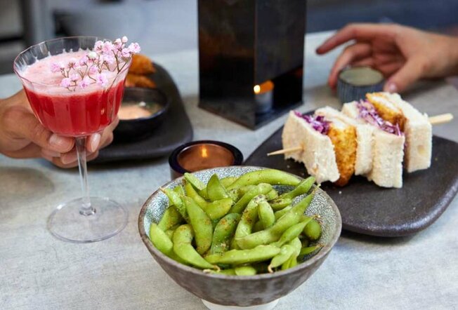 A pink cocktail next to a fried fish sandwich and a bowl of edamame