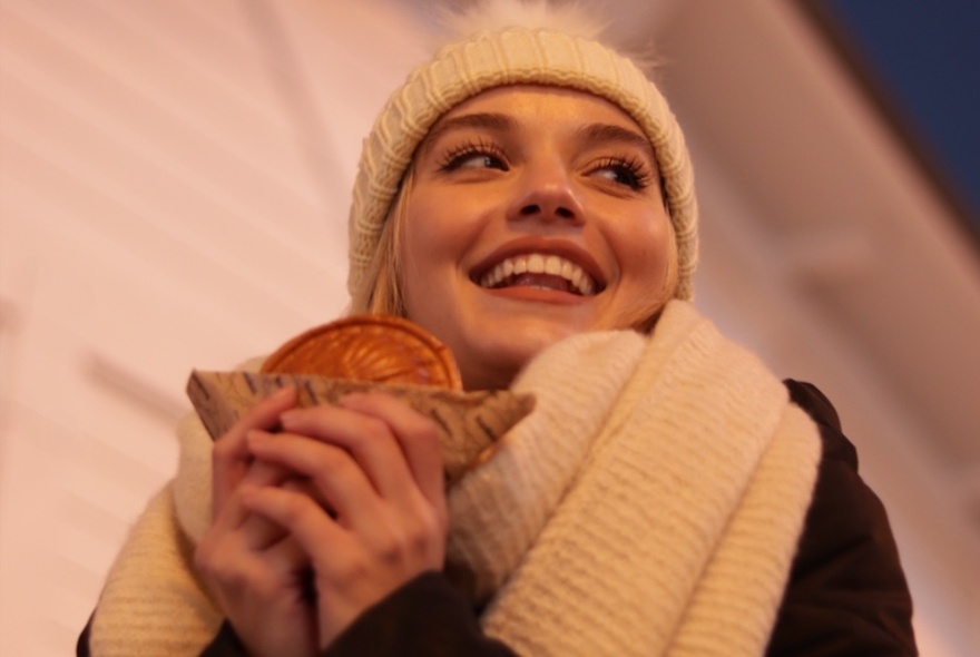 Woman in white beanie and scarf, looking to right and smiling, holding sweet in serviette, seen from slightly below.