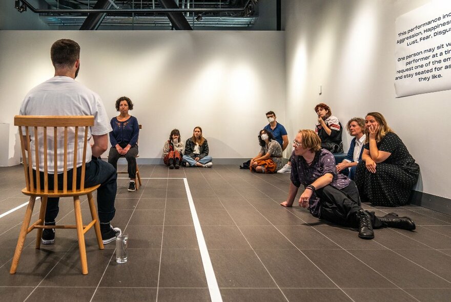 The back view of a man in a chair in a performance space with people sitting on the floor looking on. 