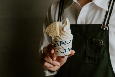 Person wearing a white shirt and a dark apron holding a takeaway cardboard cup filled with a slice of cake and soft serve cream.