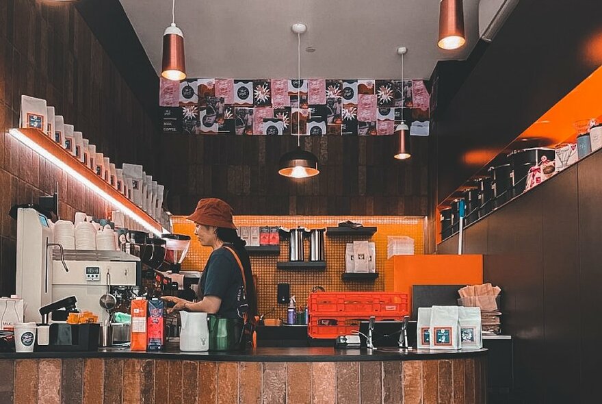 A barista making coffee behind a wooden counter in a coffee shop with orange walls and lots of coffee paraphernalia.