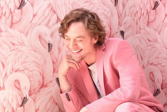 Singer Darren Hayes, smiling and with eyes closed, wearing a pink suit and posing against a wall decorated with pink flamingos.